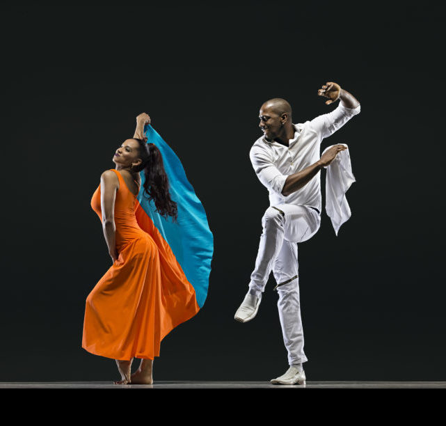 dancer in orange poses next to dancer in white smiling and lifting a knee & handkerchief