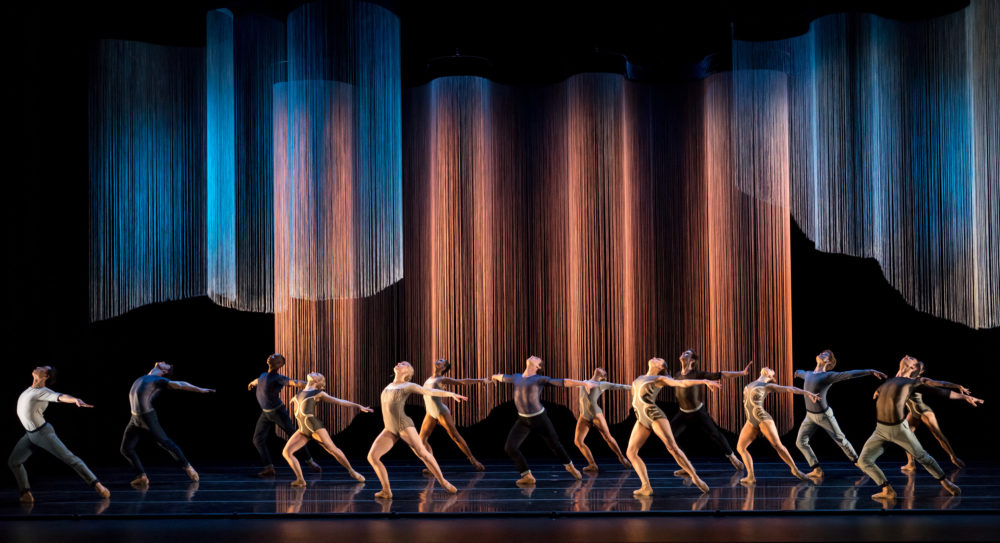 Ensemble of dancers lunge upwards on stage under colored ropes