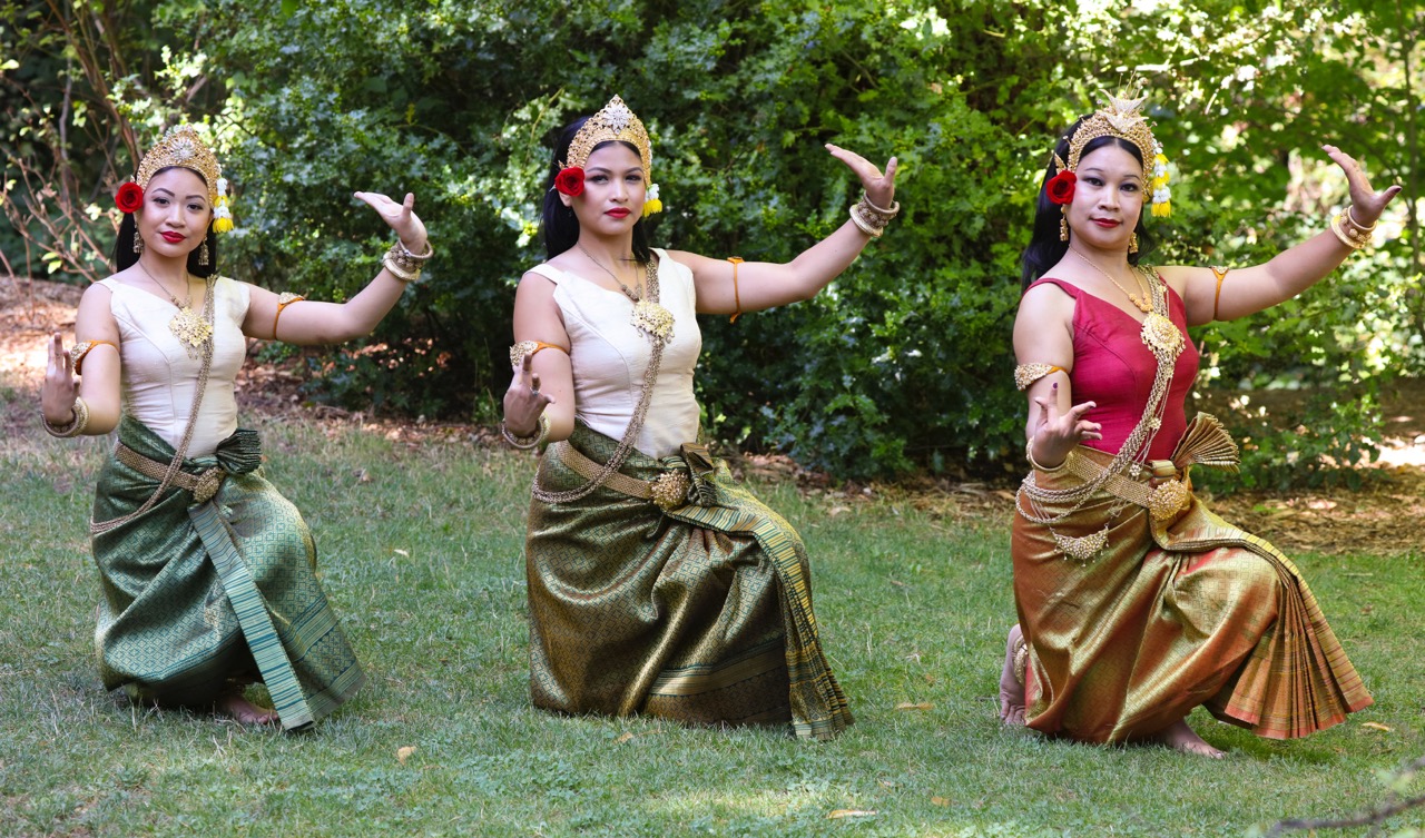 Three dancers kneeling on the grass with their arms extended and have metal headdresses