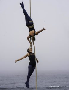 Two aerial dancers hanging from rope above the ocean.