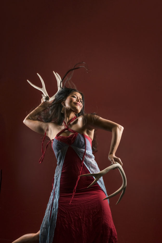 A woman holding dear antlers, one is held by her head, the other by her hip.