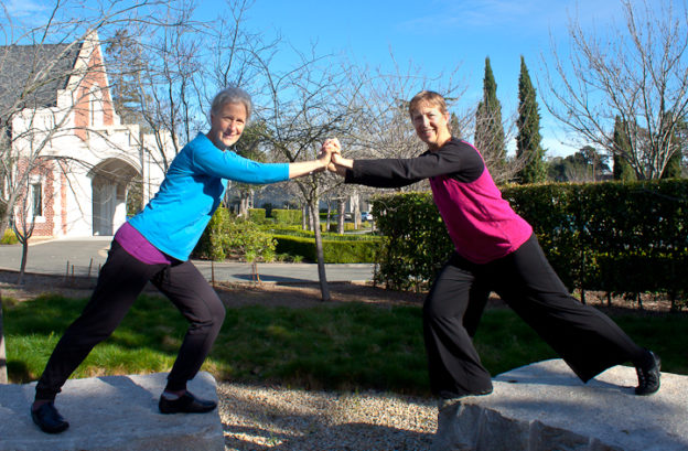 Two women are lunging on top of cemetery blocks grasping each others' hands smiling at the camera.