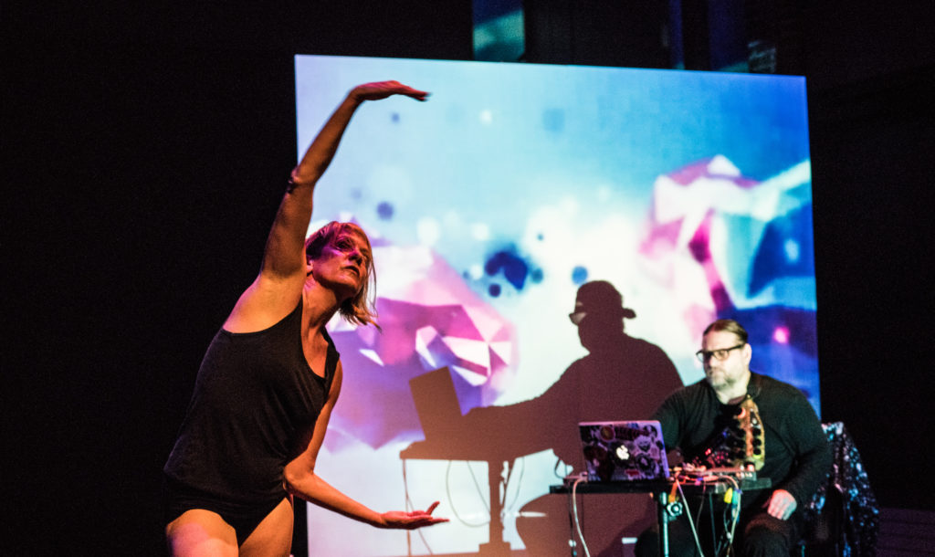 One performer making a C with her arms with a multi-colored screen and a live DJ/percussionist in the background. 