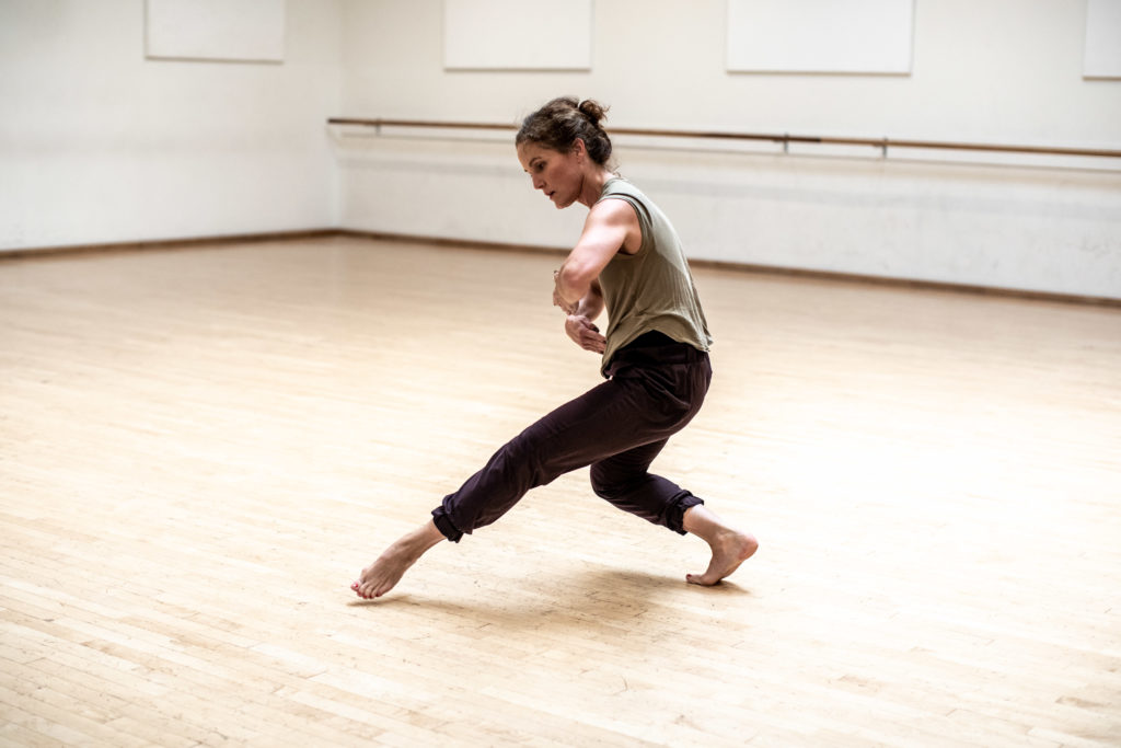 A dancer stepping forward in a deep plié with fingertips curving inwards to the chest.