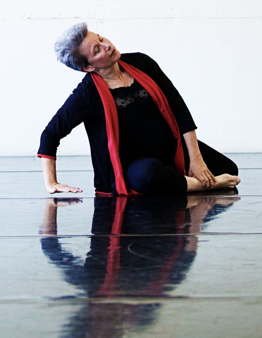 A current image of Ginny Matthews. Her lower body is lying on the floor with bent legs. One hand is on the floor holding up her upper body and the other hand is grabbing her ankle.