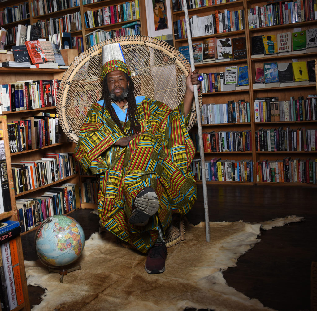 A man wearing bright multicolored robe and headdress sitting in a woven chair at a library holding a staff.