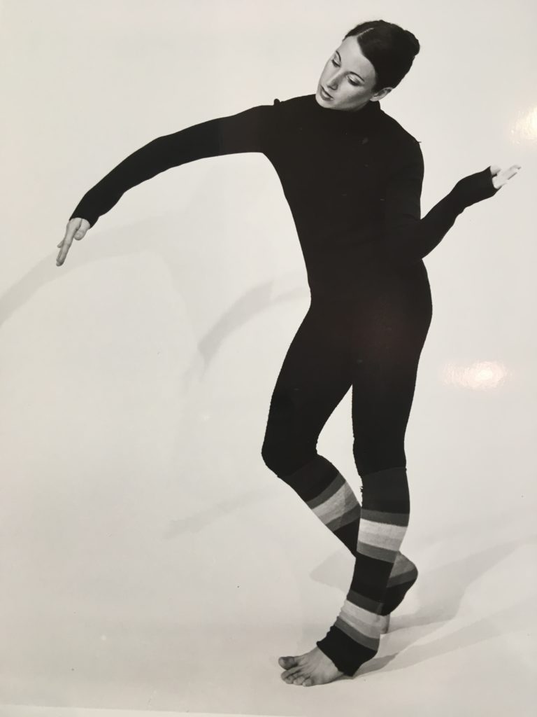 An image of Ginny Matthews from 1975. She is standing with one leg bent and her right arm in an over curve and the left arm bent at the elbow upward by her hip.