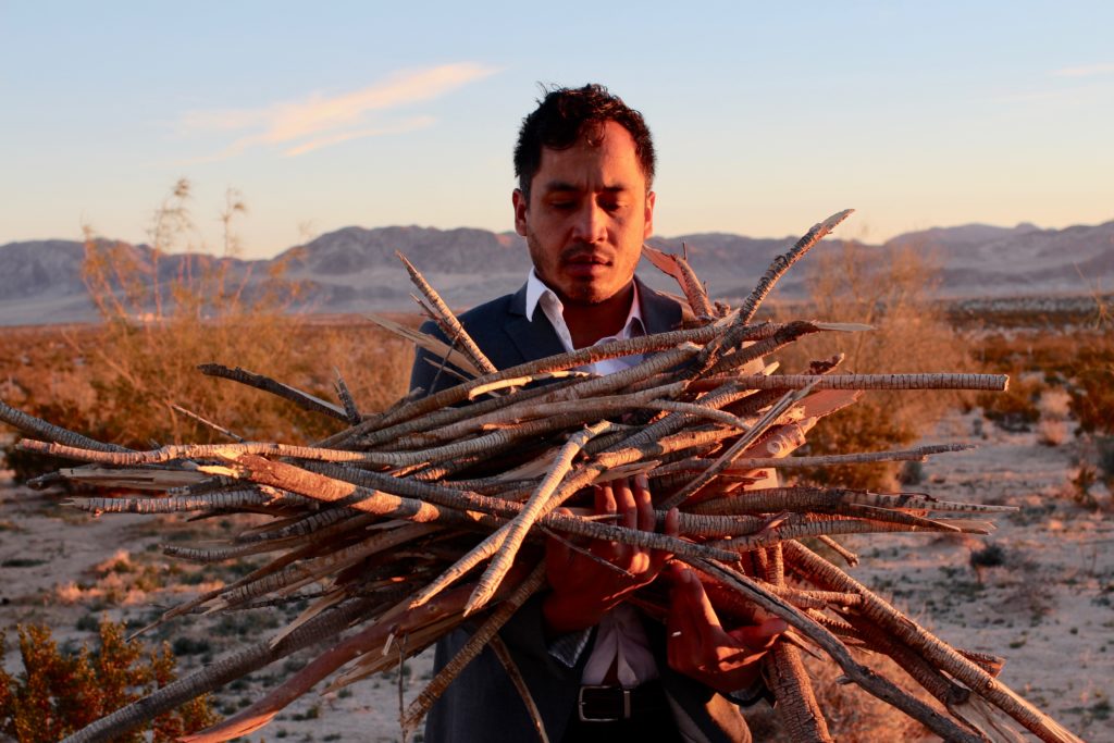 An image of Melecio Estrella holding a pile of sticks in the desert sunset.