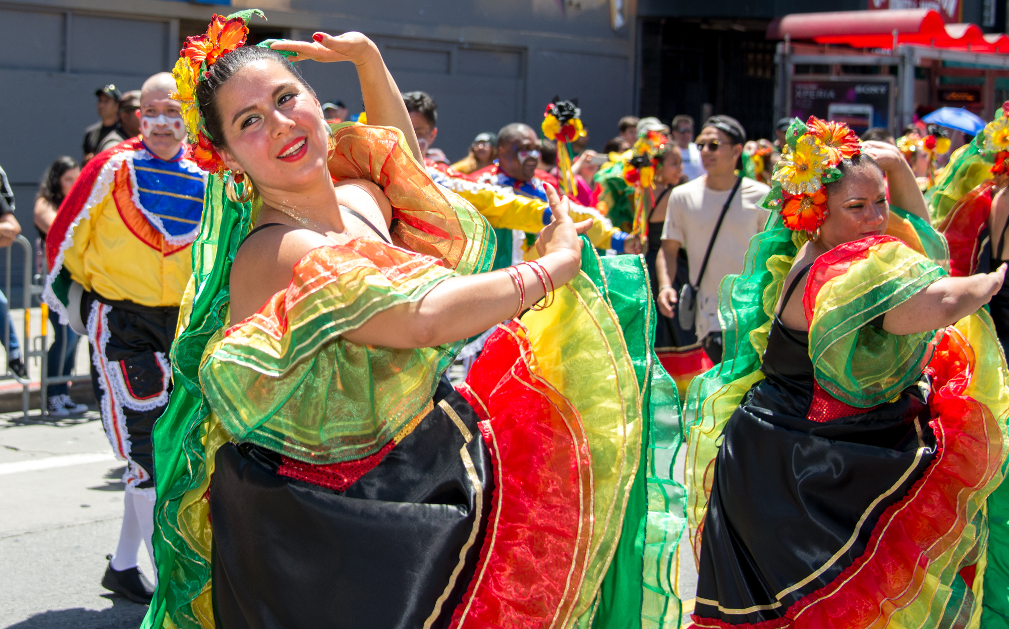 Dancers performing Columbian folkloric dance outside. They have one hand by their head with the other hand pushing outward across the chest while smiling.