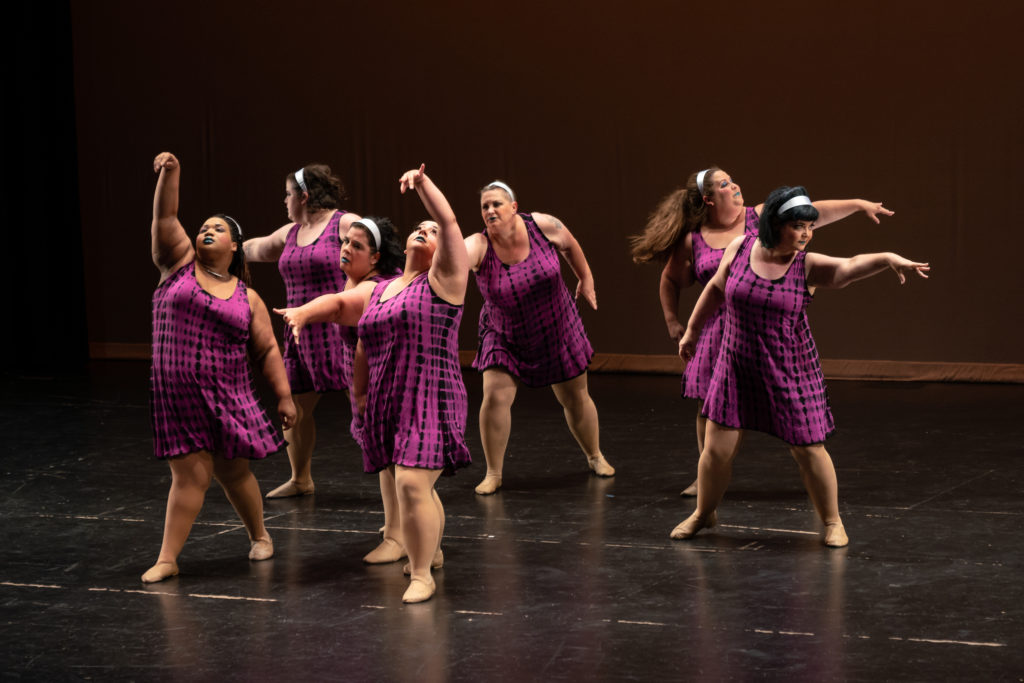 A group of six women on stage reaching in different directions.