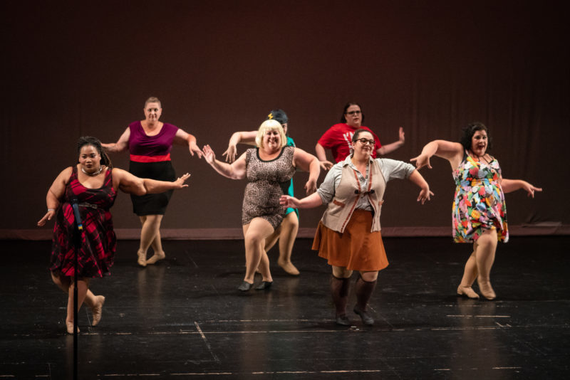 A group of seven women on stage cross stepping with wavy arms.