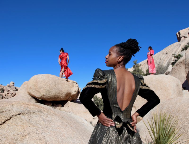 Three women standing in dresses on a rocky desert landscape.