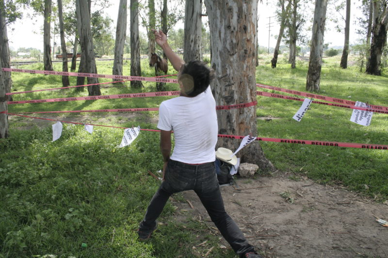 A person standing in a sparsely forested area with red caution tape and paper with writings on them. The person is slightly leaning back with one arm up, back facing the camera.