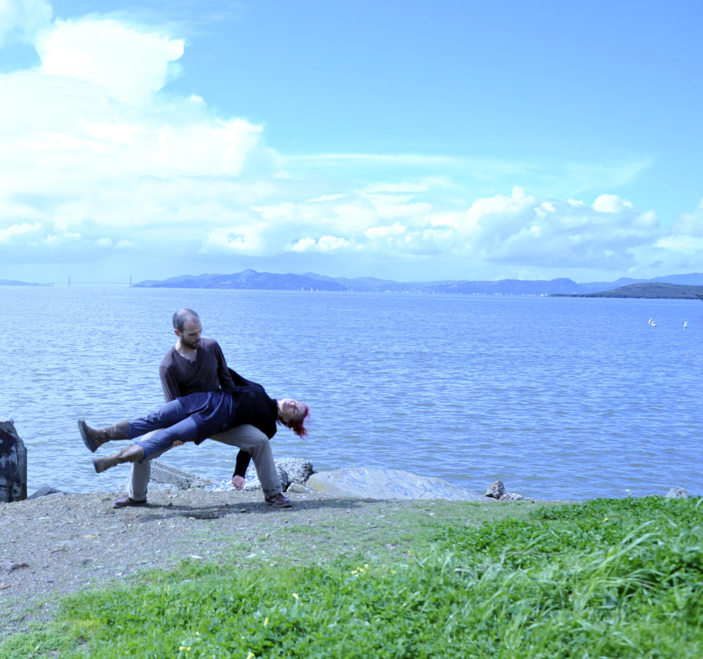 Two people near the coast line. A man is supporting the whole body of a woman on his legs. He is holding her in a squat as she lies flat and stiff.