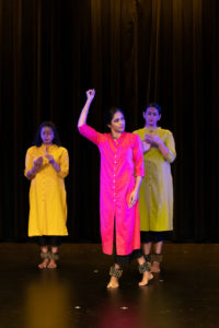 A bharatanatyam dancer with an arm up walks in front of two other dancers focused on their hands