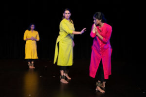 Three bharatanatyam dancers in a diagonal with the front person looking back at another whose arm is extended towards them