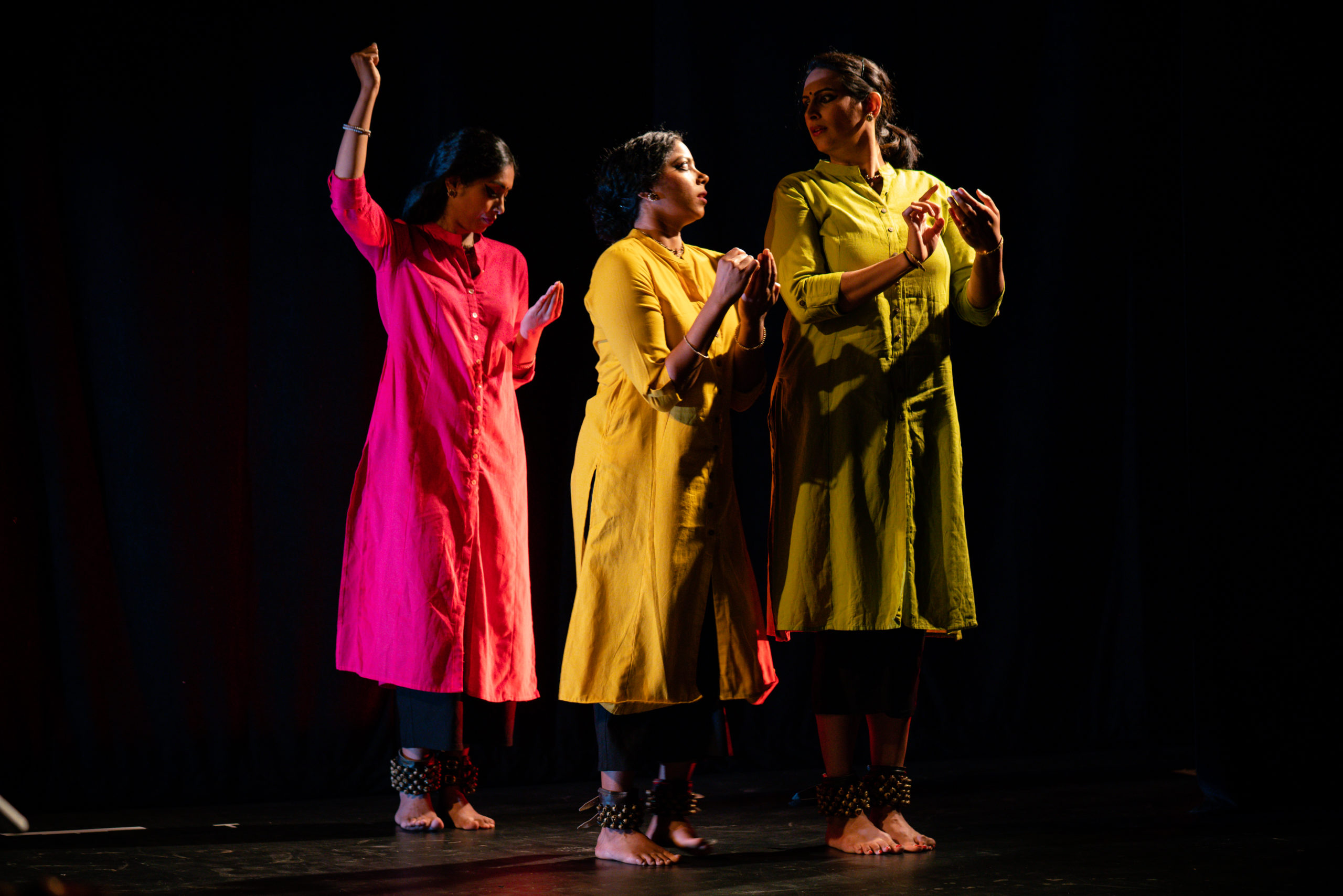 Three dancers in bright pink, yellow, and green stand close together onstage in a bharatanatyam performance
