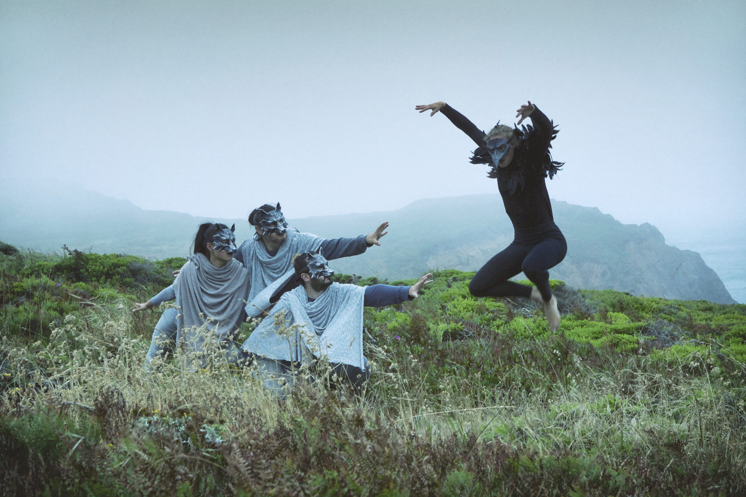 On a seaside cliff, three dancers in wolf masks and light gray clothing focus on a leaping dancer in all black with a crow mask