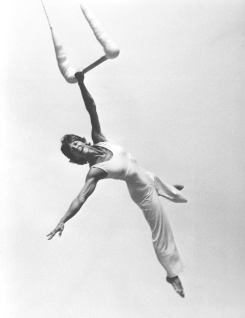 Black & white photo of Terry Sendgraff flying in the air and hanging onto a trapeze with one arm