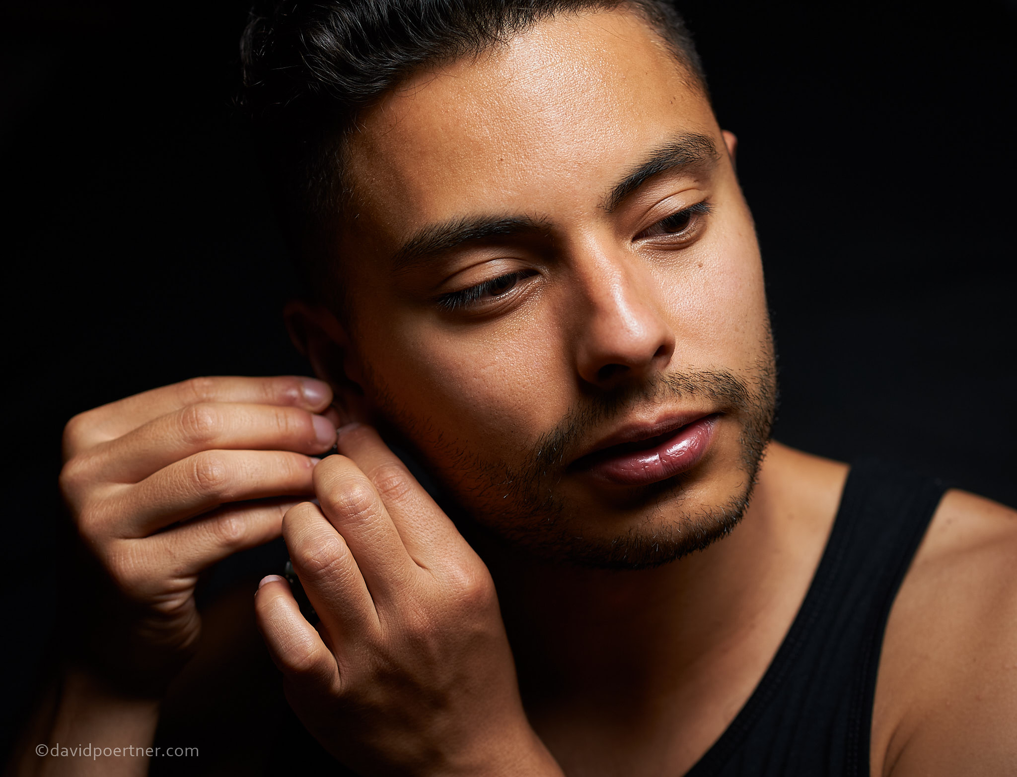 headshot of Juan Urbina showing Juan in a black tank top, Juan's face with eyes looking down and off to the side and hands near their right ear