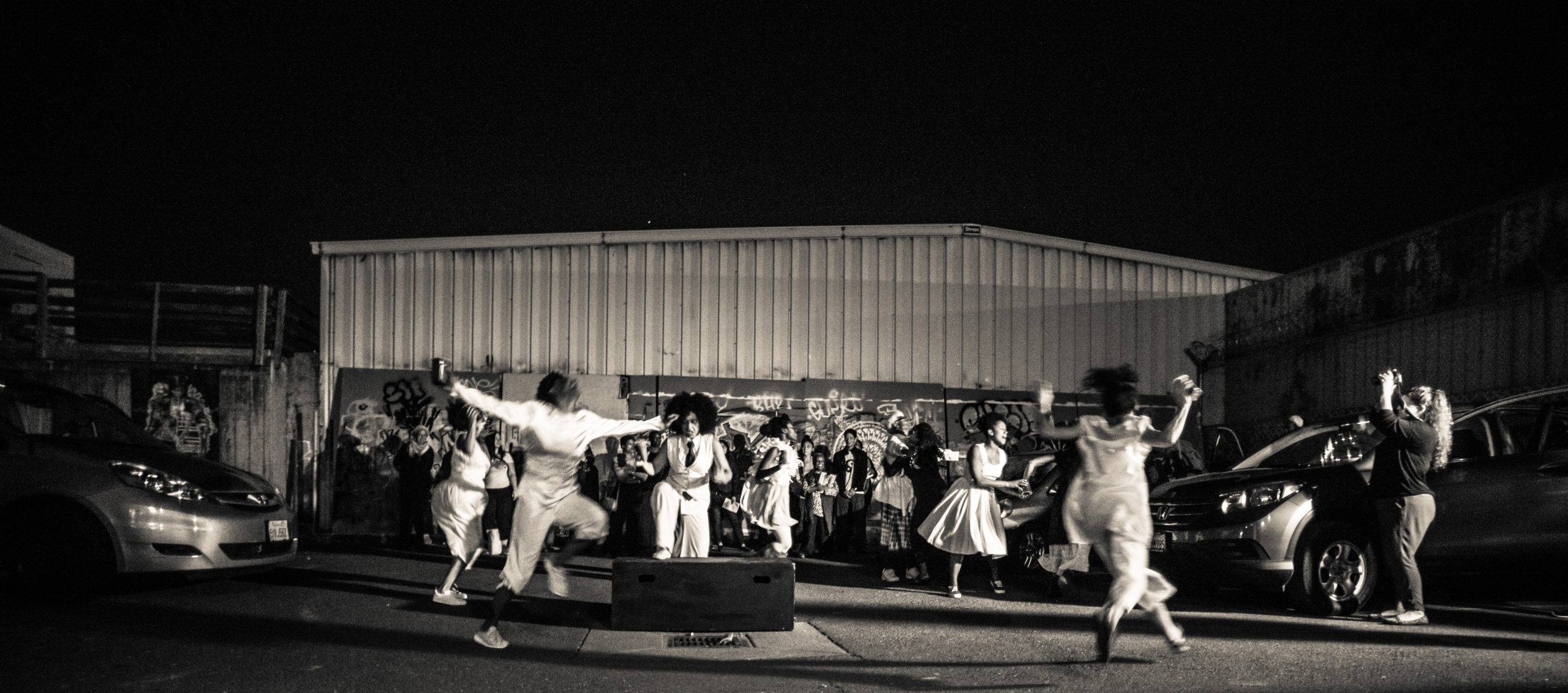 [Image description: Group of performers clothed in different variations of white dresses, pants, shirts, and skirts dancing in a parking lot between several cars. One woman with big curly hair, in a white pantsuit with a black tie steps up onto a box in the middle of the circle.]