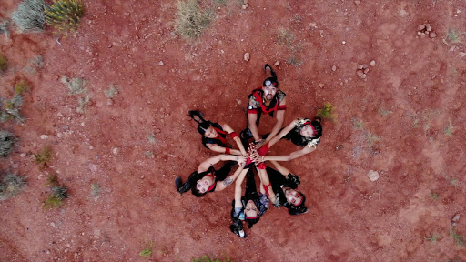 A group of dancers form a circle and reach to the middle with their arms while looking upwards towards the camera