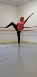 A woman standing with one leg bent and the opposite arm reaching away on a diagonal in a dance studio.
