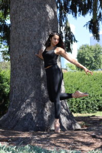 Emma Garber poses on pointe by a tree, looking down and bending one foot behind her