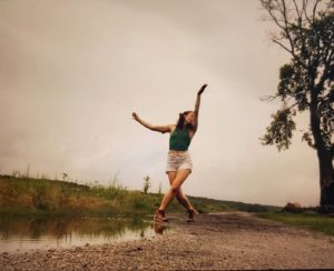 A current photo of the author Julia Davidson. She is dancing outside on a gravel road near a tree and a puddle.