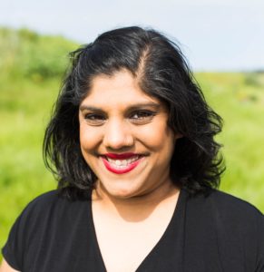 A brown-skinned desi person stands against a bright green, out of focus background smiling toward the camera.
