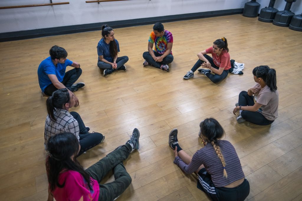 People sit in a circle on the brown wooden floor of a white-walled dance studio.