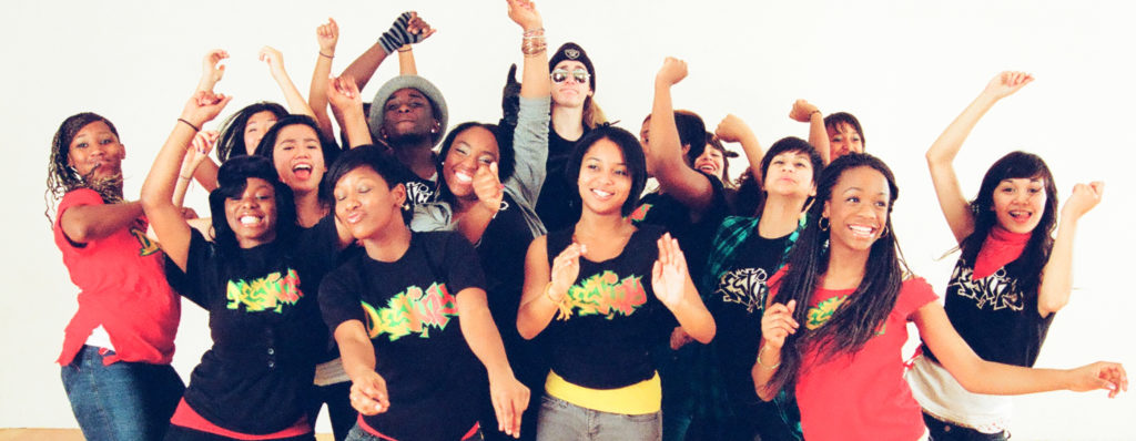 Group of 13 dancers with arms up and faces beaming with happiness, wearing black and red t-shirts with colorful graffiti writing.