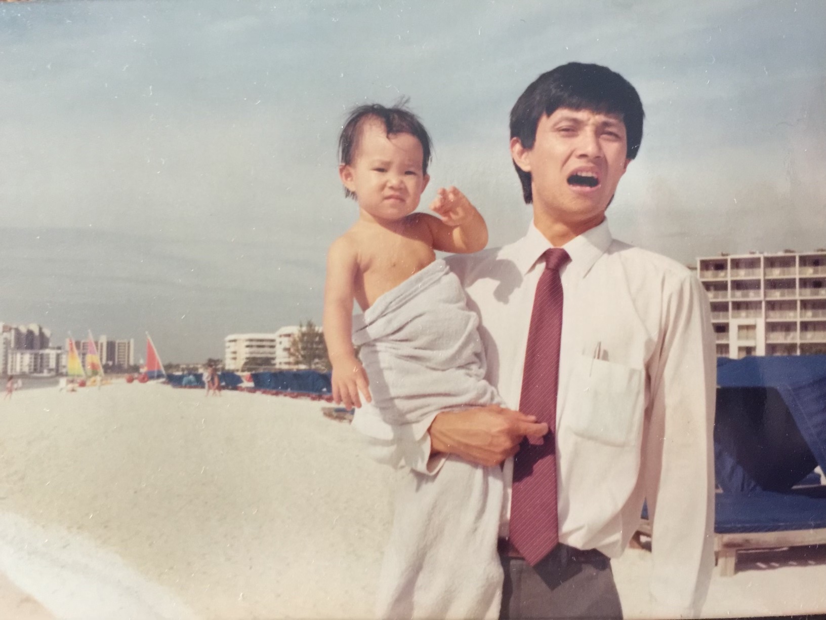 A photo of a young Chinese father holding his baby daughter half-wrapped in a towel, with a white sand beach in the background.