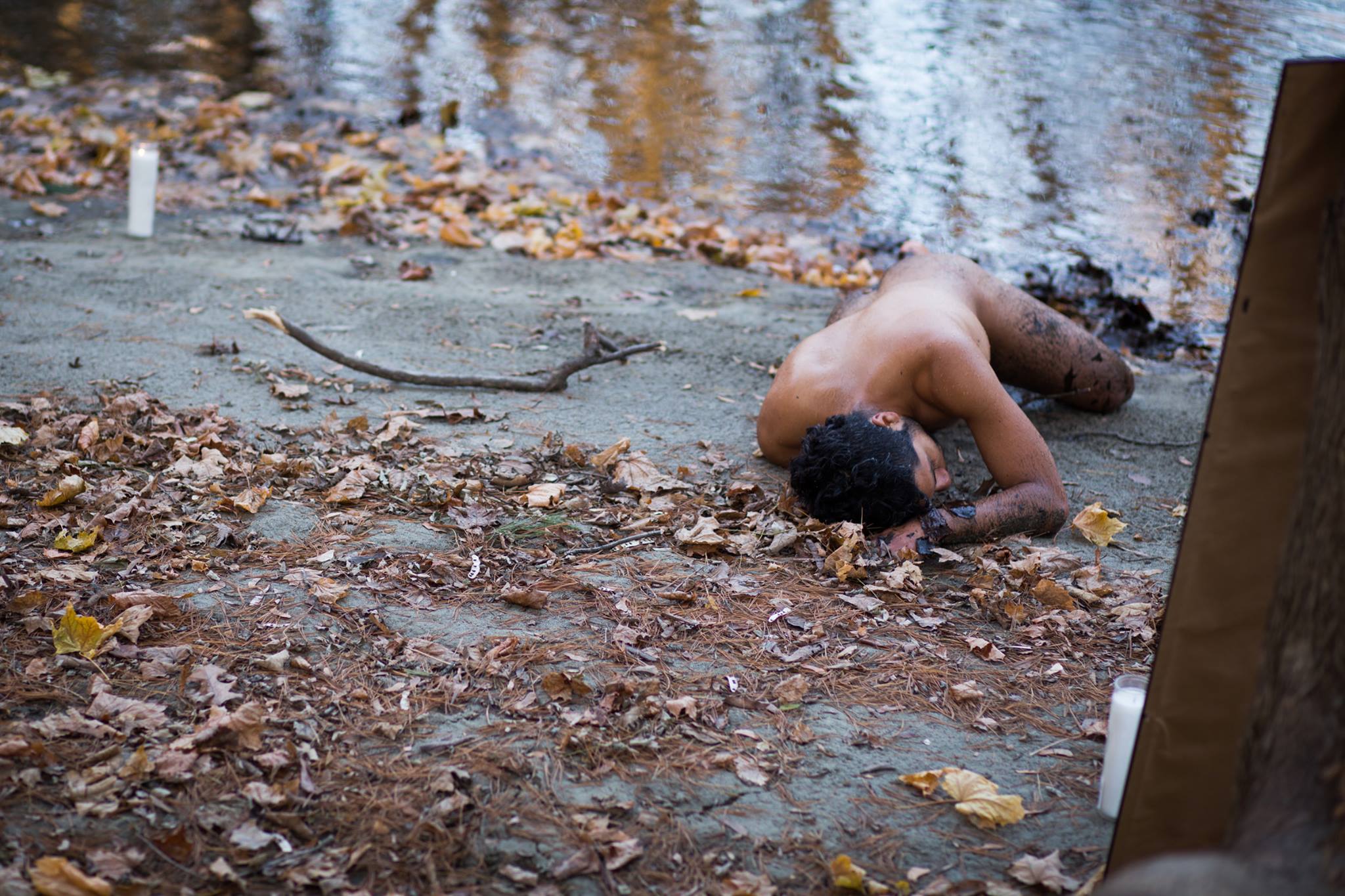 Estrellx is laying naked, face down, spiritually surrendering their body along a riverbank with their Brown body covered in debris