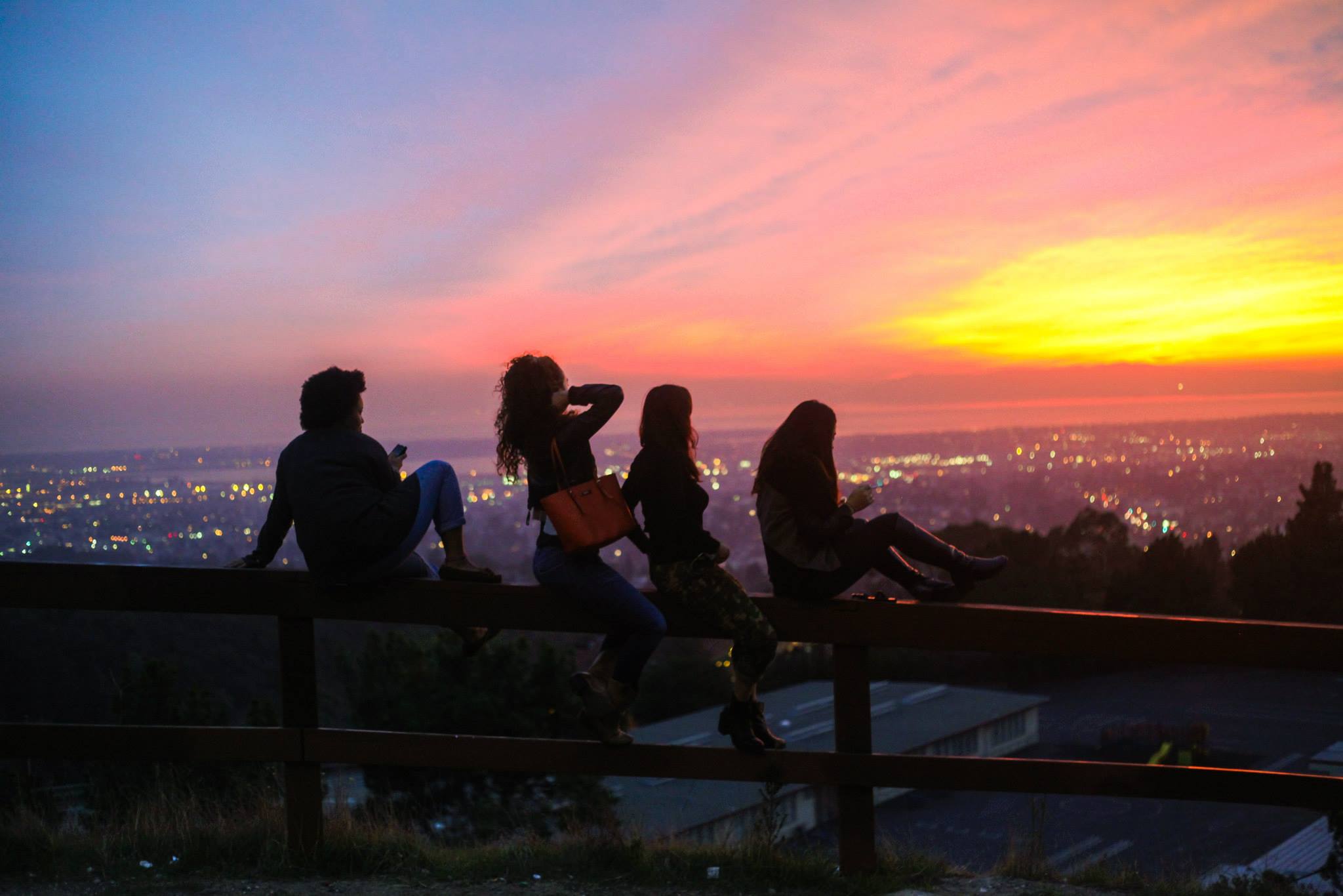 Four teenage girls sit on top of a wooden fence facing the sunset