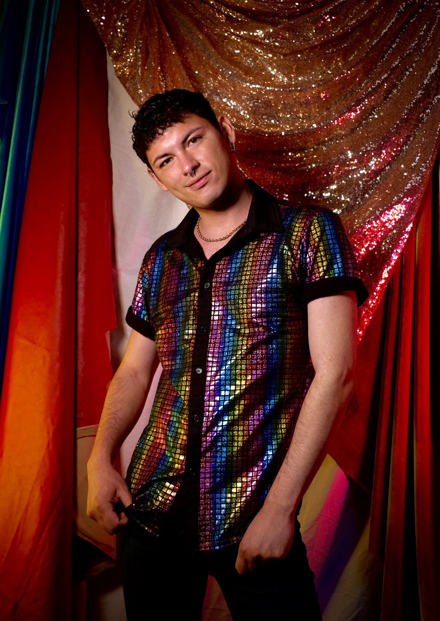 Jesse stands in front of a red sequin curtain while wearing a checkered rainbow button up shirt.