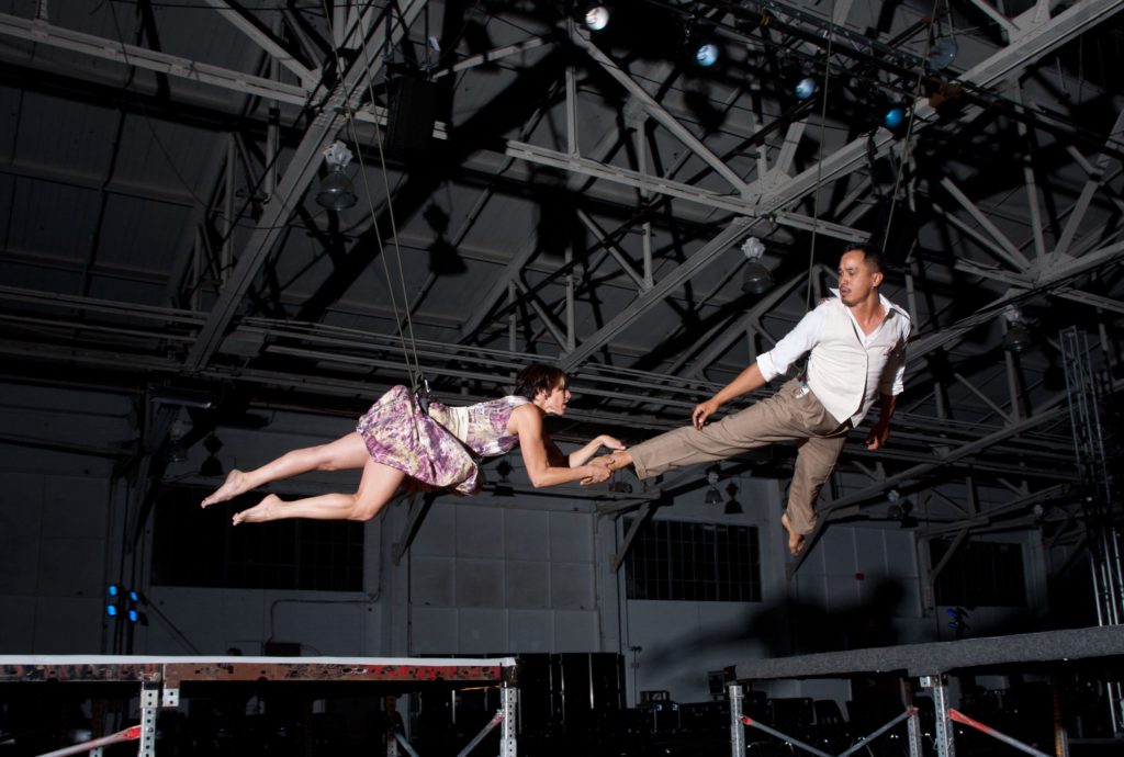 Two dancers suspended by ropes flying in duet. One dancer has hold of the other's ankle.