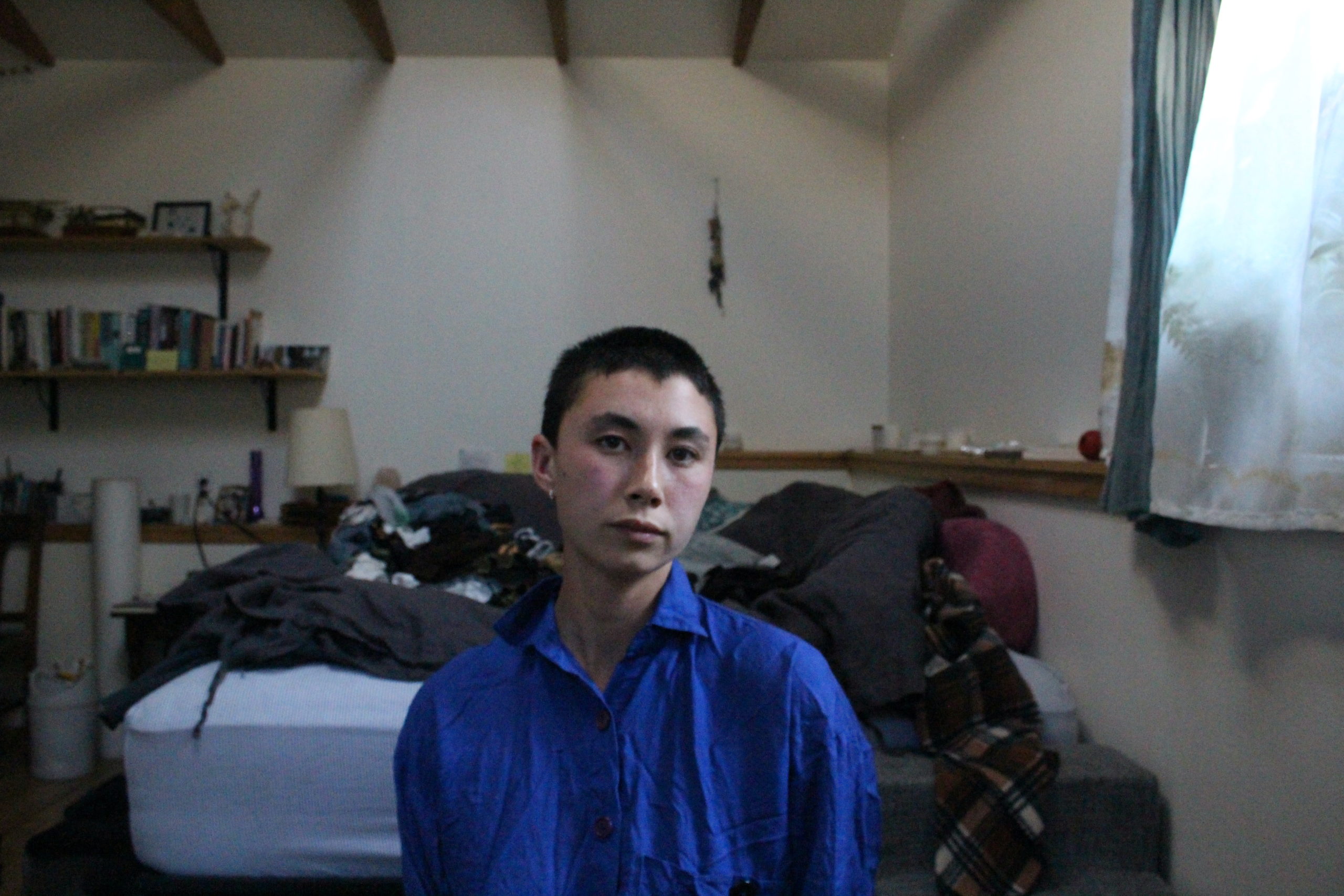 Portrait of person in a blue shirt in their room, positioned in front of a bed that is full of blankets.