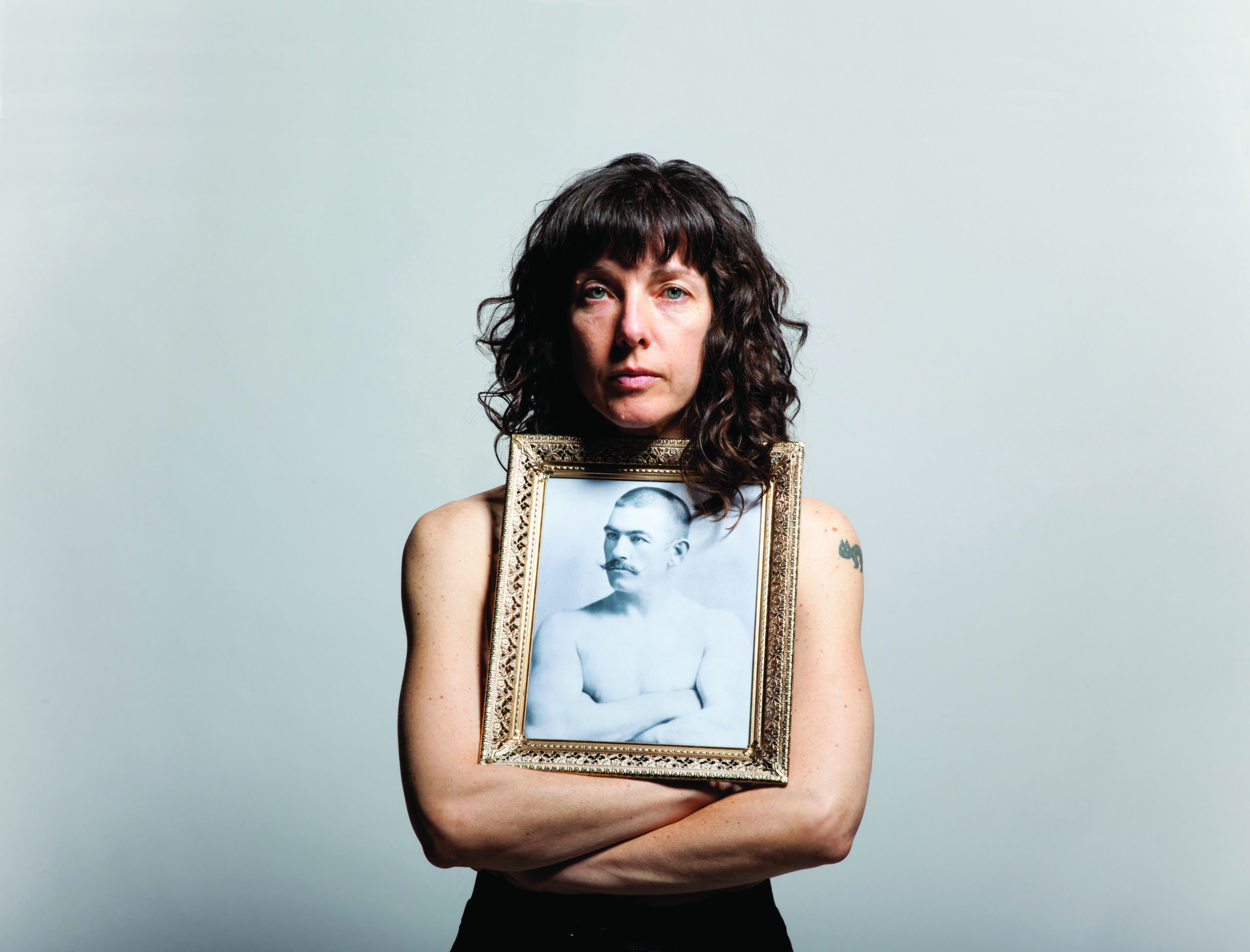 Chris Black, a white woman with long dark hair, faces the camera holding a picture of a man with a shaved head in the same pose