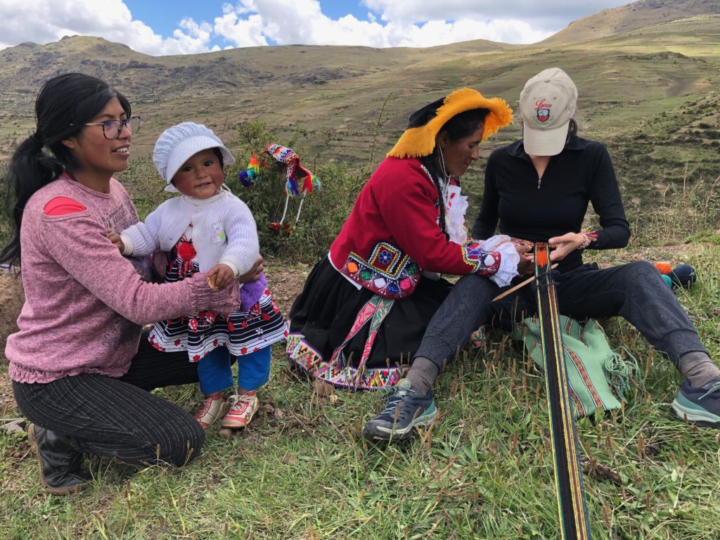A young Peruvian mother holds her smiling baby while the Peruvian grandmother helps an American learn how to weave from the waist.