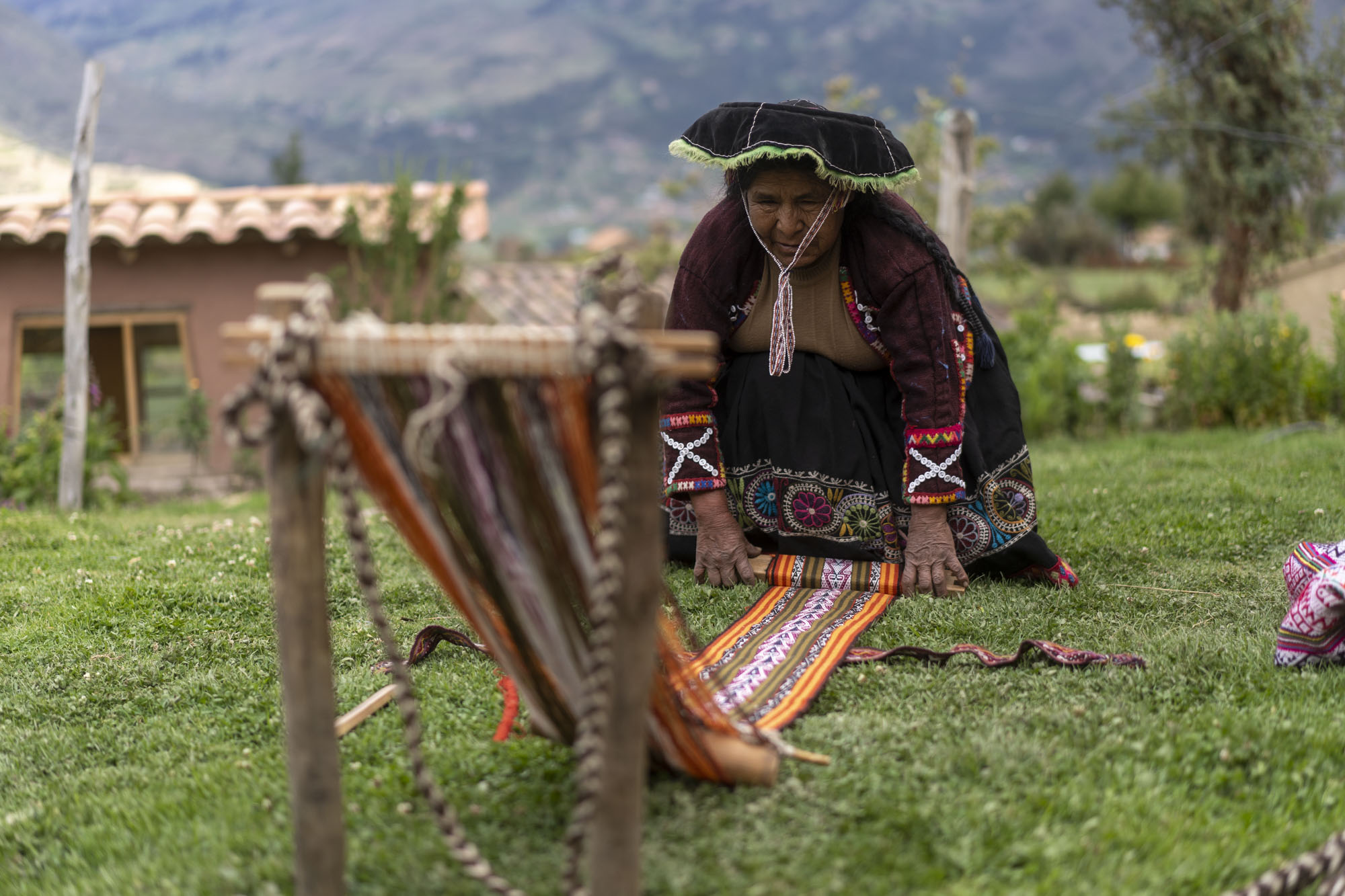 An older Peruvian woman with a broad black hat unrolls her tapestry on the ground in front of the Peruvian Andes mountains.