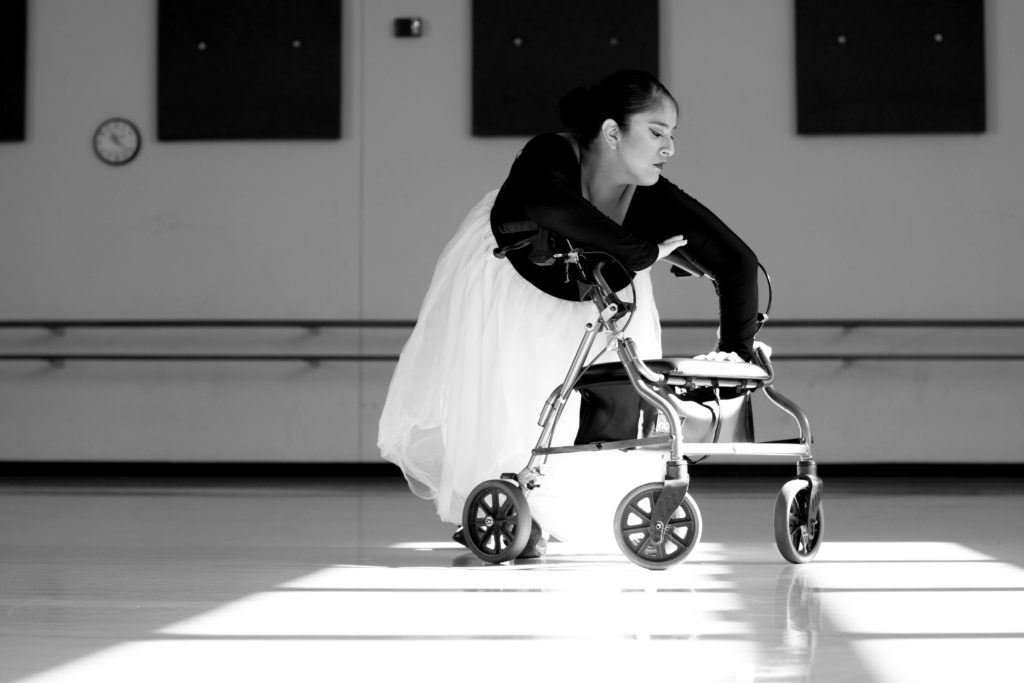 A black & white photo of Vanessa Hernández Cruz gracefully dancing with her walker. She is wearing a ballet dress that has a tulle skirt.