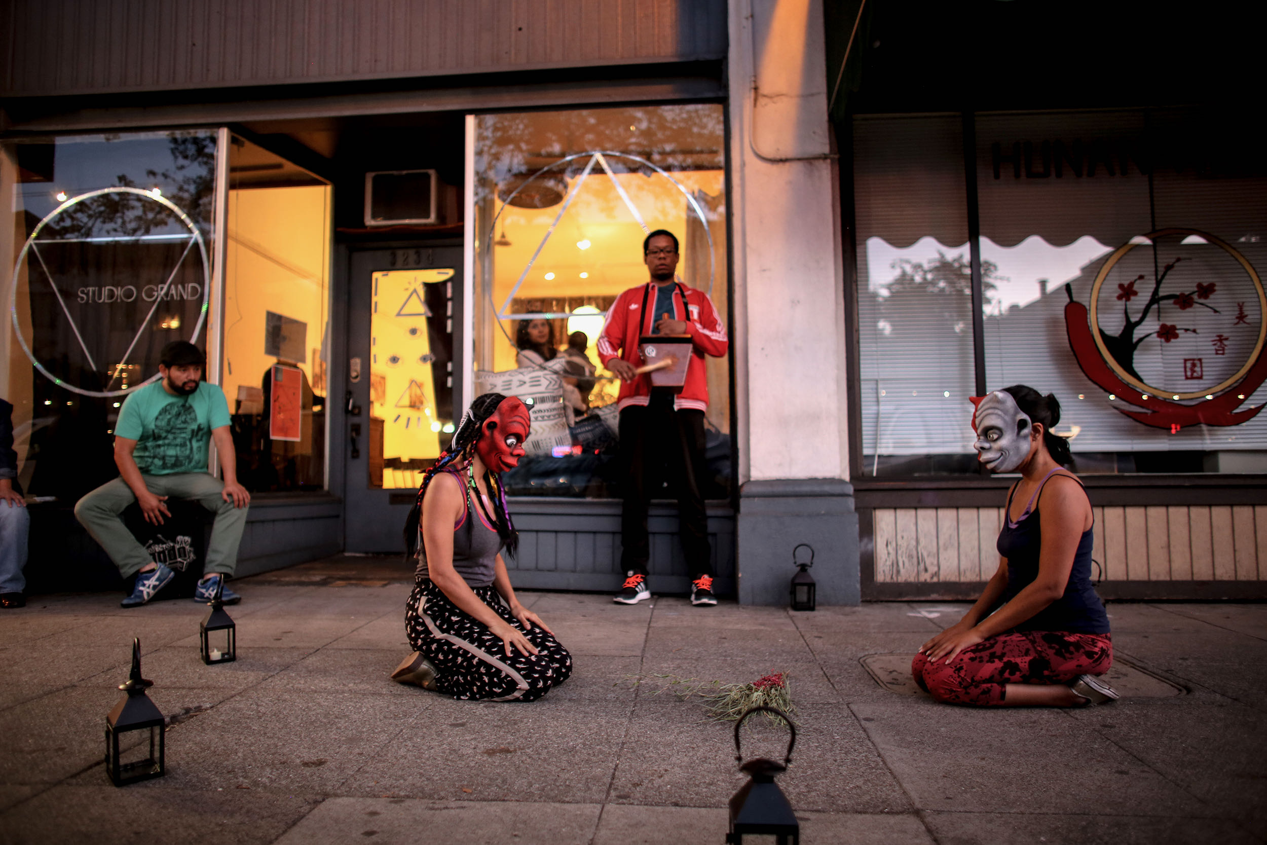 Two dancers in masks kneel on the sidewalk. Behind them a woman holding a baby, a man with a cajita, and another with a Cajon.