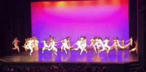 Large group of dancers in white costumes against a pink and purple backdrop. The dancers are jumping in unison.