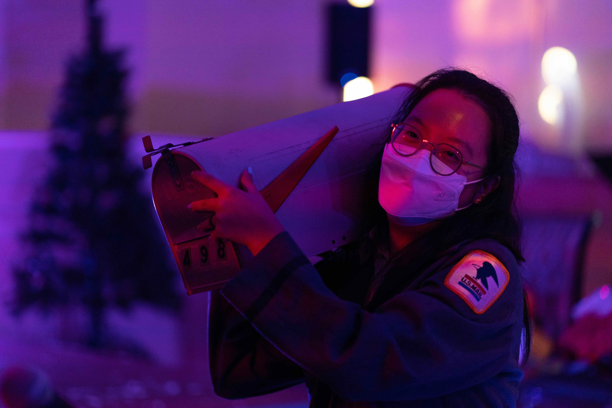 Haley Baek is in a postal worker uniform, glasses, and a white mask and carries a mailbox on her shoulder, looking delighted.