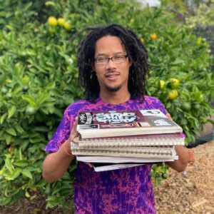 A Brown Man wearing a purple shirt with dreadlocks and glasses holding a sketchbook