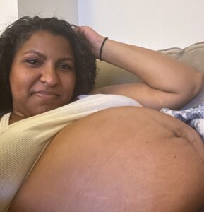 Danielle sits back on a couch with an arm behind her head and pregnant belly exposed.