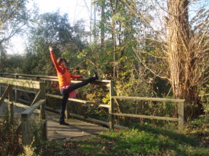A middle-aged white woman in a puffy orange vest does a dancerly pose on a footbridge in the woods.
