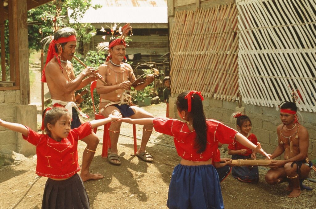 Two dancers in red tops and blue skirts dance as two male elders play music. A father and daughter crouch to play music, too.