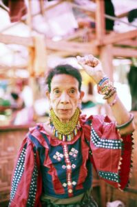 A female elder in center frame raises her left hand in a fist while adorned with traditional clothing and accessories.